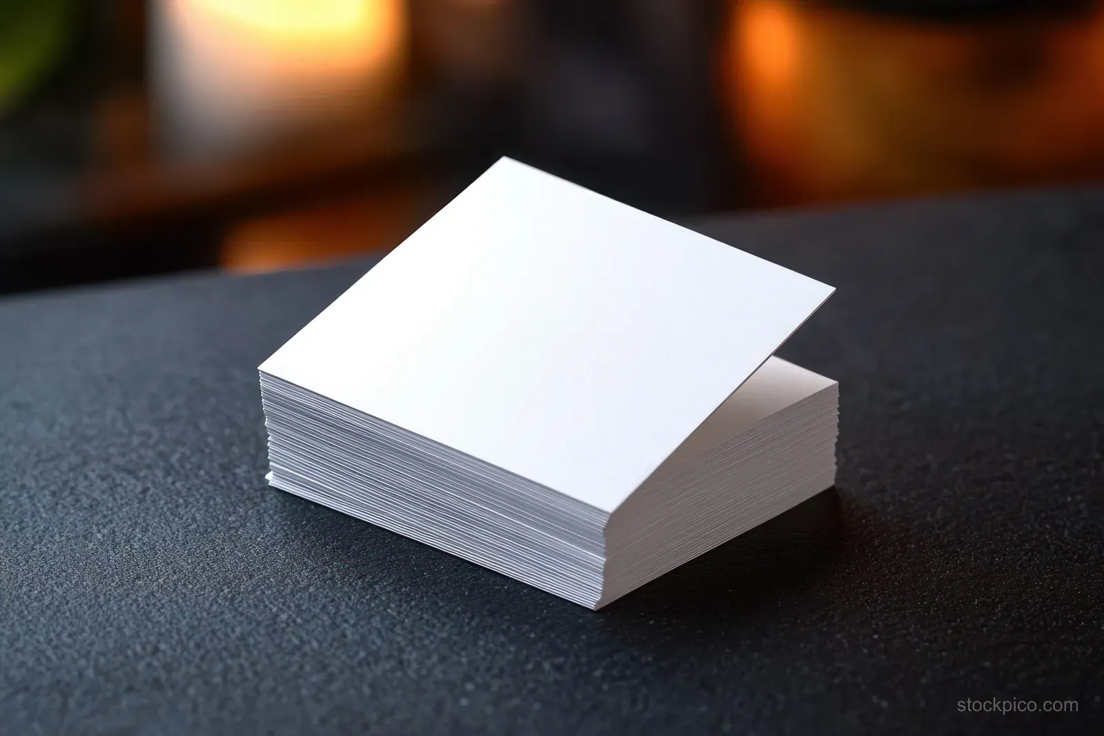 A small stack of blank white square papers sits on a dark textured desk with warm blurred lights in the background.