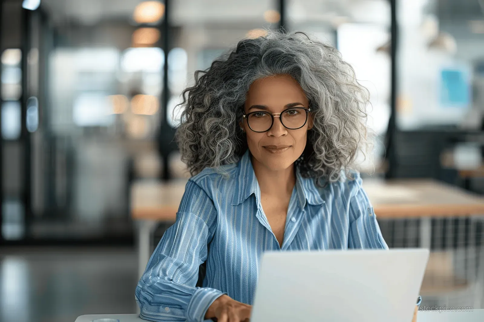 A middle-aged businesswoman with glasses and curly gray hair works on a laptop at a desk in a modern glass-walled office.