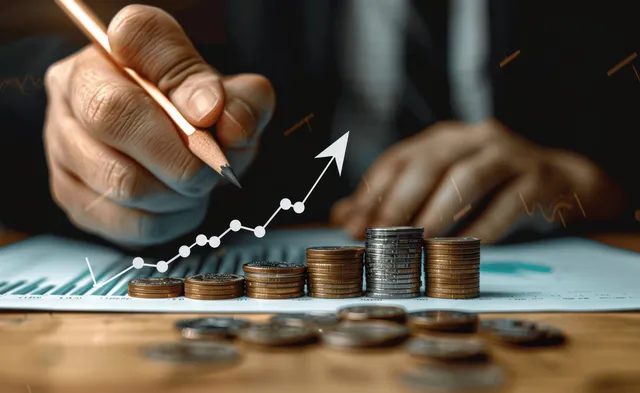 A hand holding a pen draws an upward arrow on a finance chart beside stacked coins and a ruler.