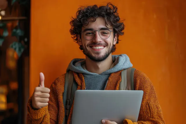 Happy young man with curly hair and glasses giving thumbs up while holding laptop