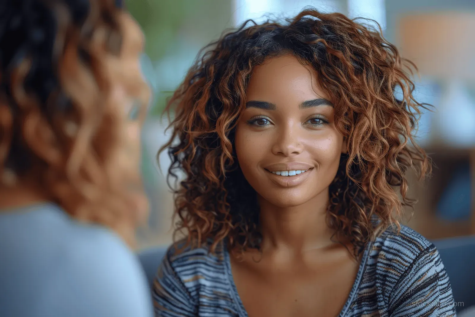 A woman with curly hair smiles at the camera while wearing a striped top in a softly lit indoor setting.