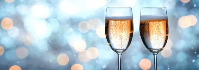 Two champagne flutes clink in front of snowy winter bokeh lights with golden sparkling highlights.