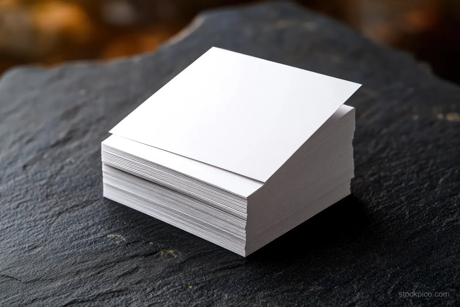 Stack of blank white square cards placed on a dark textured stone surface with warm blurred background.