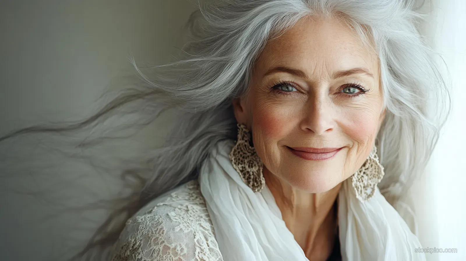 Smiling elderly woman with flowing white hair wearing lace earrings and a white lace shawl in soft indoor light.