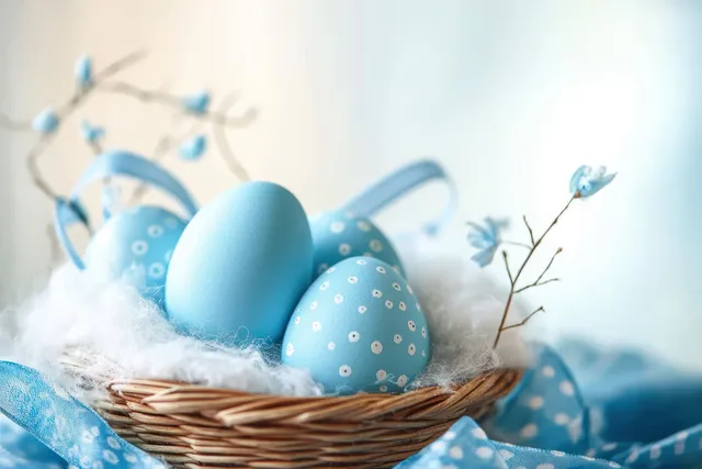 Blue decorated Easter eggs in a wicker basket resting on white fluff with small blue blossoms.