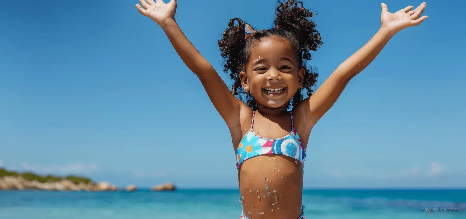 A joyful African American girl in colorful swimwear splashes with arms raised on a sunny beach under a blue sky.