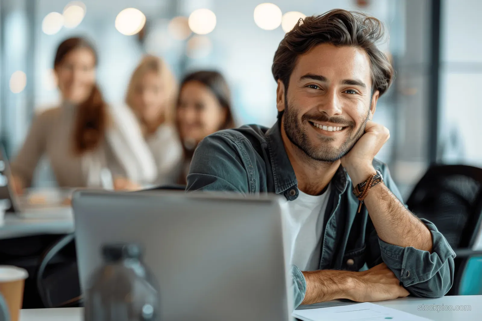 Thoughtful man rests his chin on his hand at a laptop in a bright office meeting room with coworkers blurred behind.