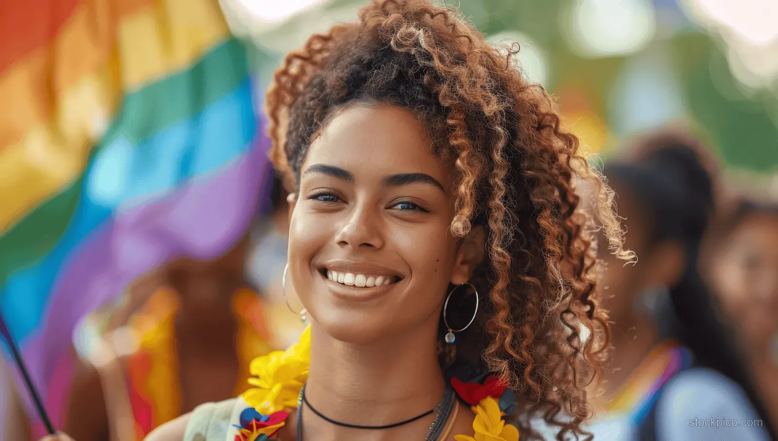 Curly-haired young woman marches at a Pride parade holding a rainbow flag amid a cheering street crowd.