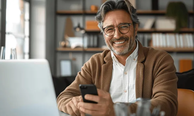 Smiling middle-aged business manager using smartphone and laptop at modern office desk