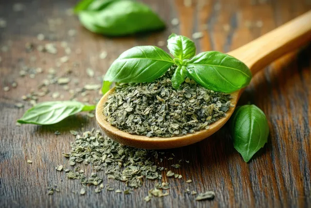 Dried herb flakes in a wooden spoon with fresh basil leaves on a rustic wooden table.