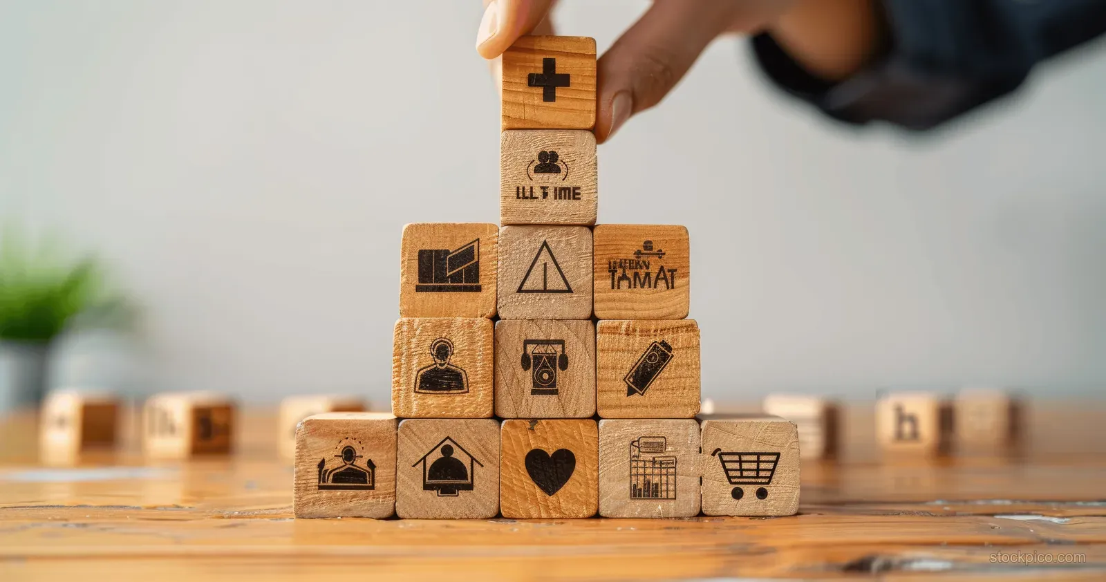A hand stacks wooden blocks forming a pyramid of employee benefit icons including insurance and vacation.