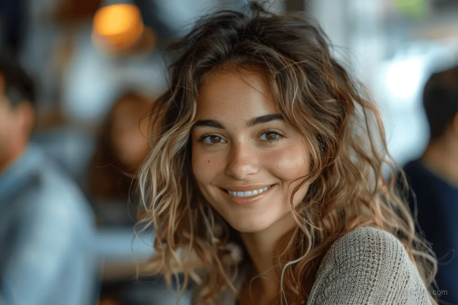 A smiling woman in an office cafe with warm bokeh lights and blurred coworkers behind her.