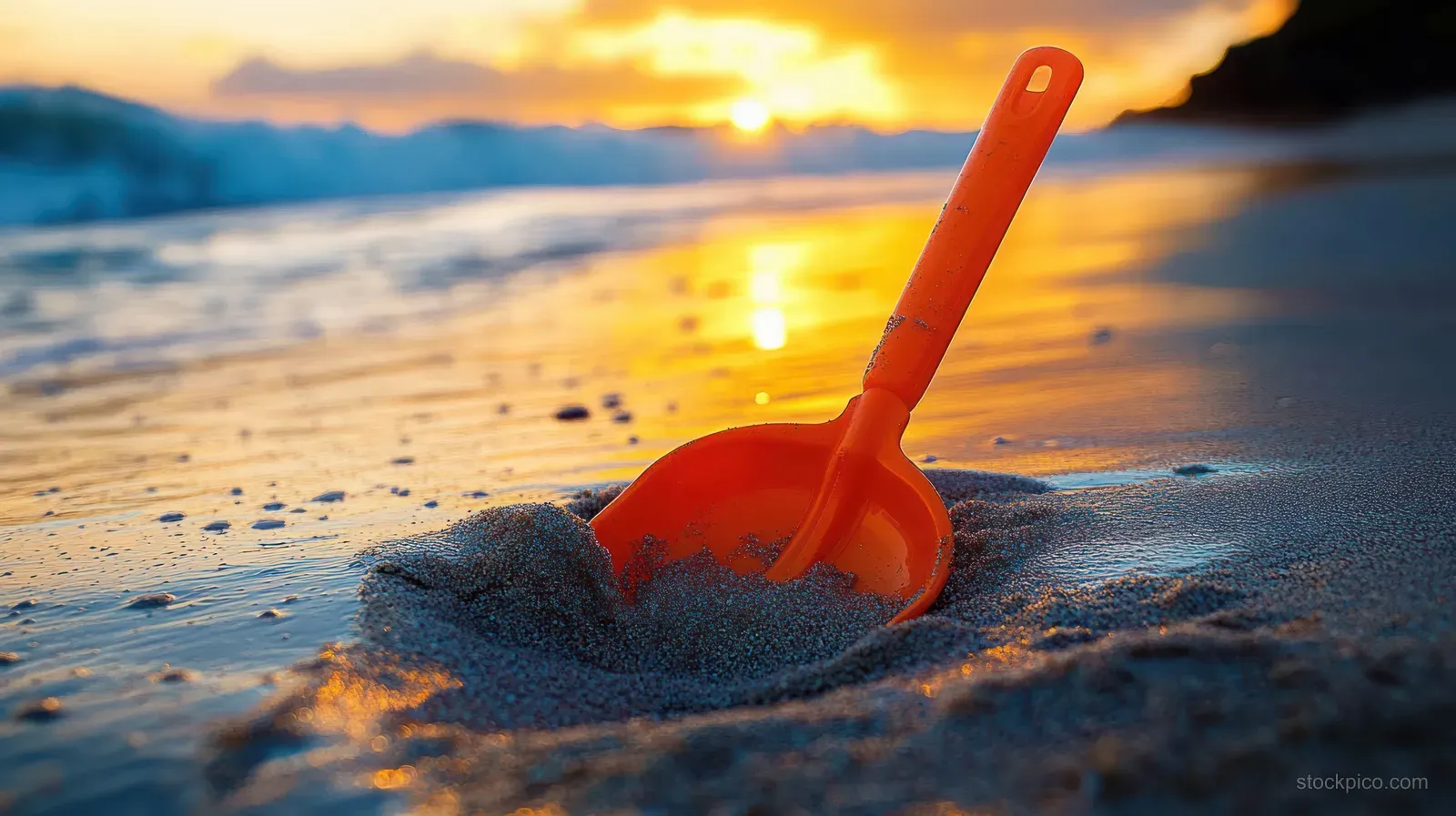 Orange toy shovel half-buried in wet sand on a beach with sunset reflections and soft ocean waves.