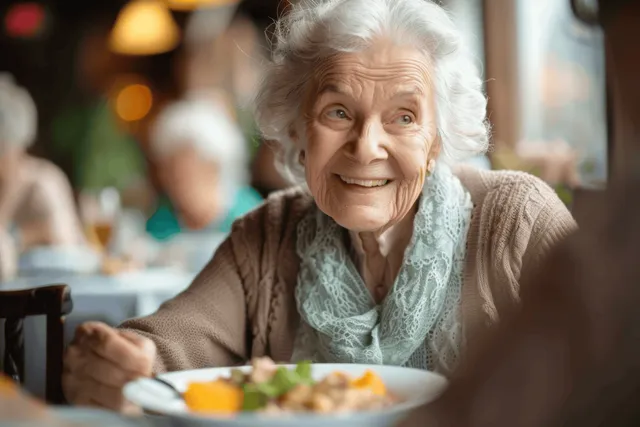 An elderly woman smiles at a dining table in a cozy restaurant while eating a meal.