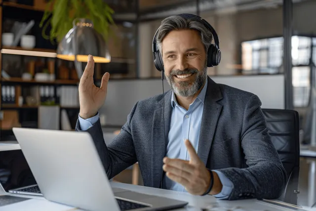 A cheerful businessman in a headset engages in a video call from a modern office, using a laptop for remote collaboration.