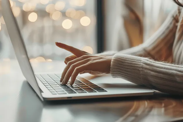 Woman’s hands typing on a laptop at an office desk with soft lights blurred behind.
