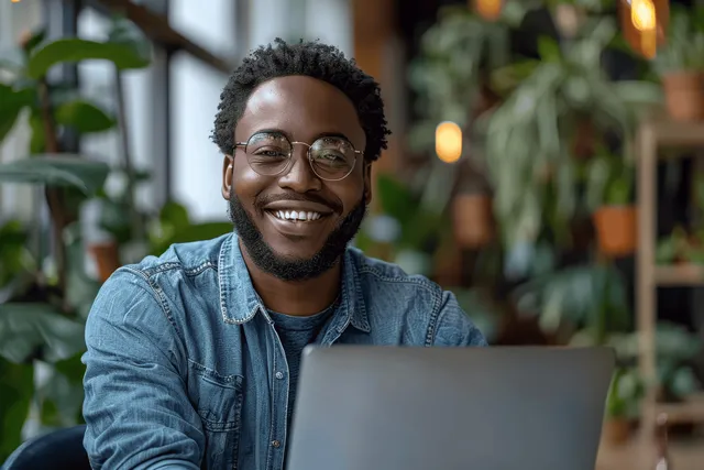 Cheerful man with glasses sits at a laptop and writes notes in a modern home office surrounded by plants.