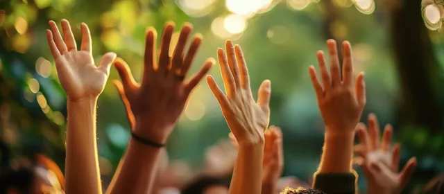 Multiple diverse people raise their hands high outdoors under warm sunlight with green trees in soft focus.