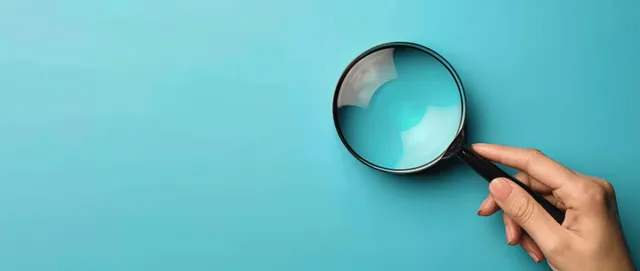 A hand holds a magnifying glass over a blue background, with a reflective lens surface.