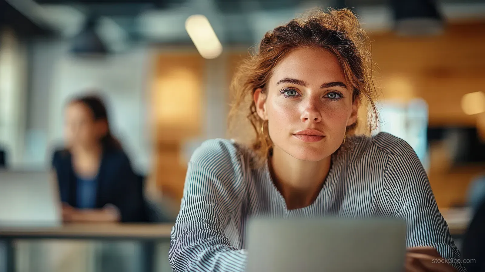 A young Hispanic woman types on a laptop at an office desk while coworkers remain softly blurred behind her.