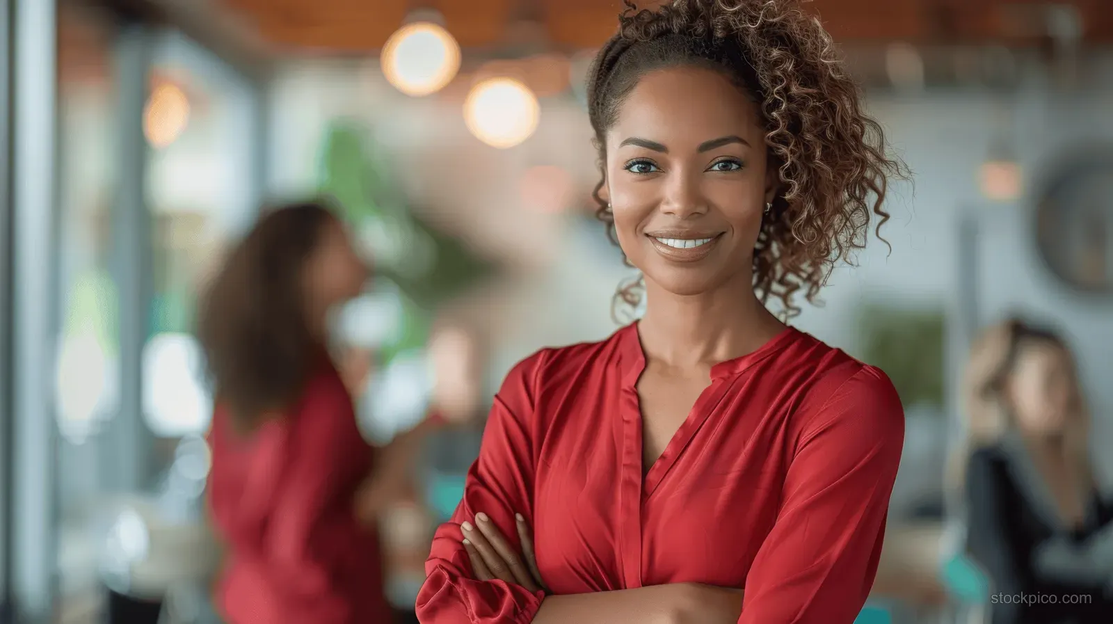 A smiling African American businesswoman in a red blouse stands in a modern office with coworkers blurred in the background.