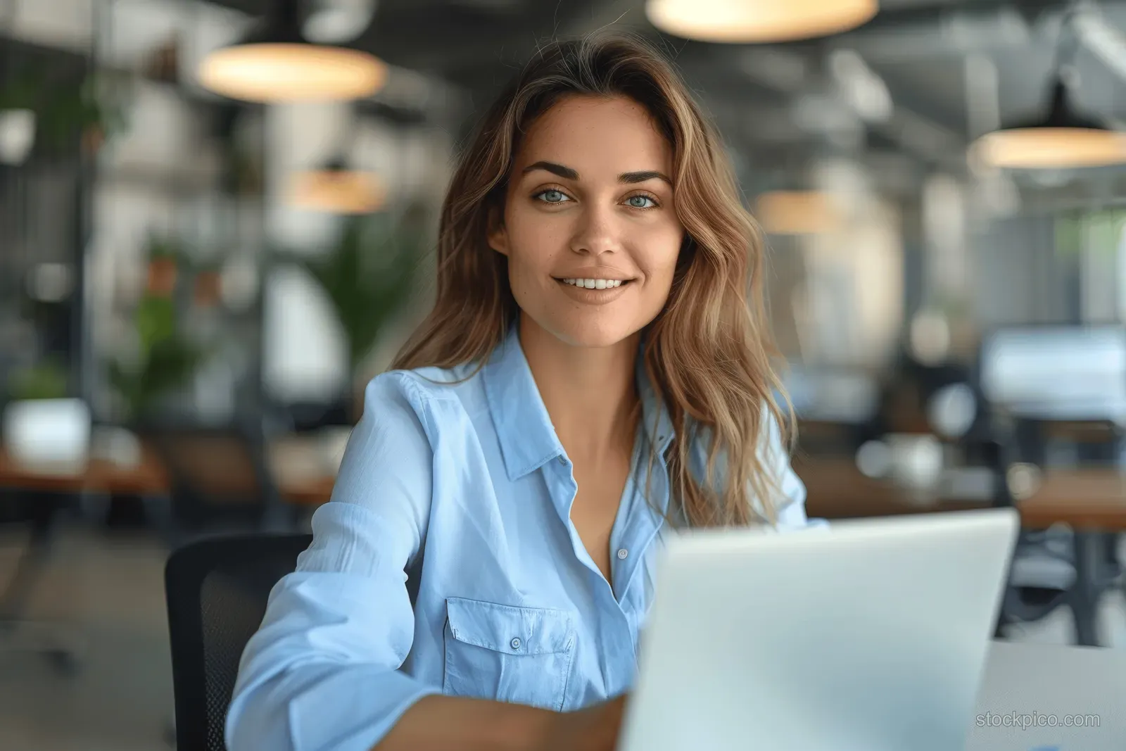 Smiling middle aged businesswoman in blue shirt typing on a laptop at a modern office desk with blurred background.