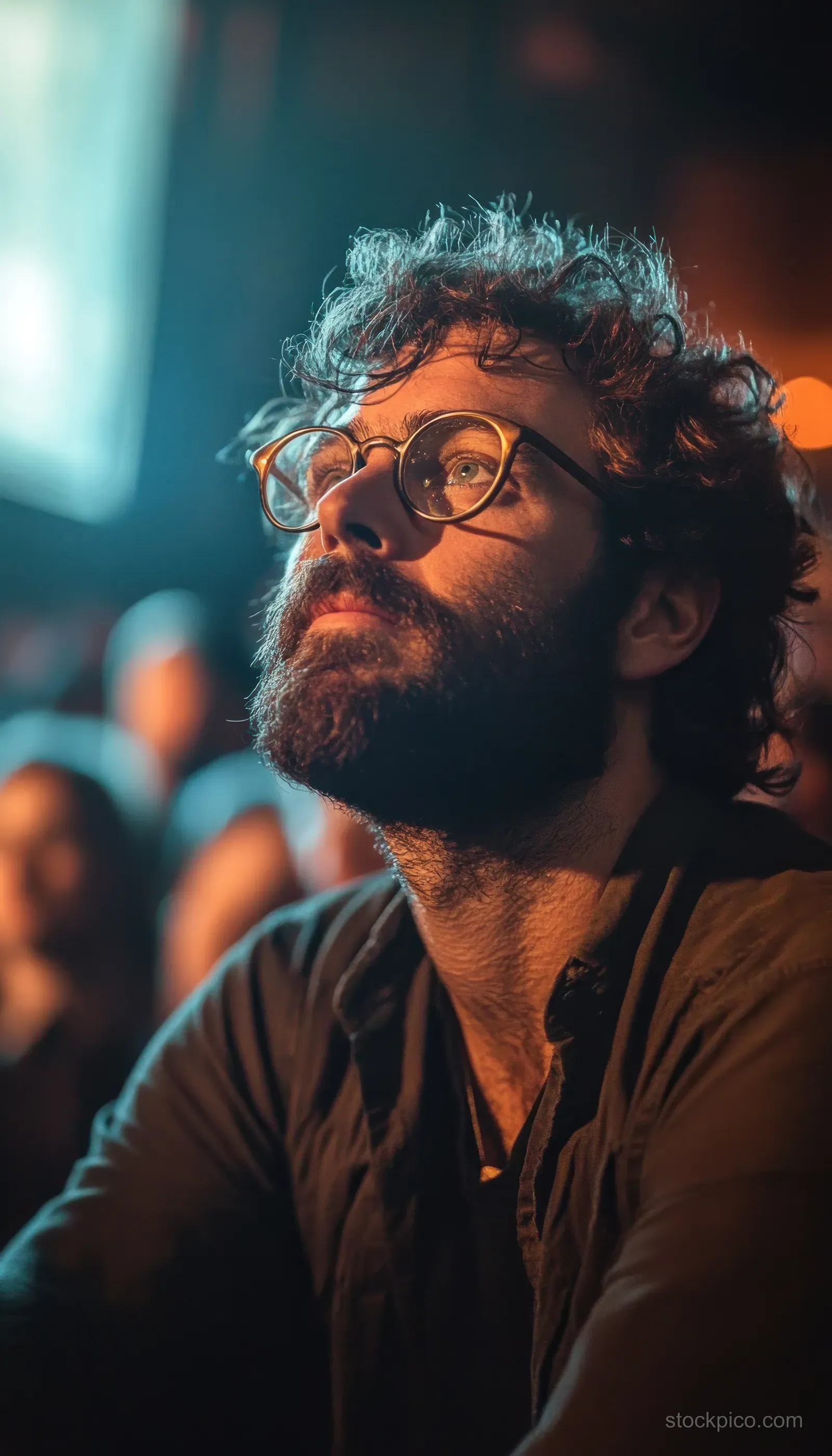 Young bearded man wearing glasses looks upward in warm indoor light while a blurred crowd fills the background.