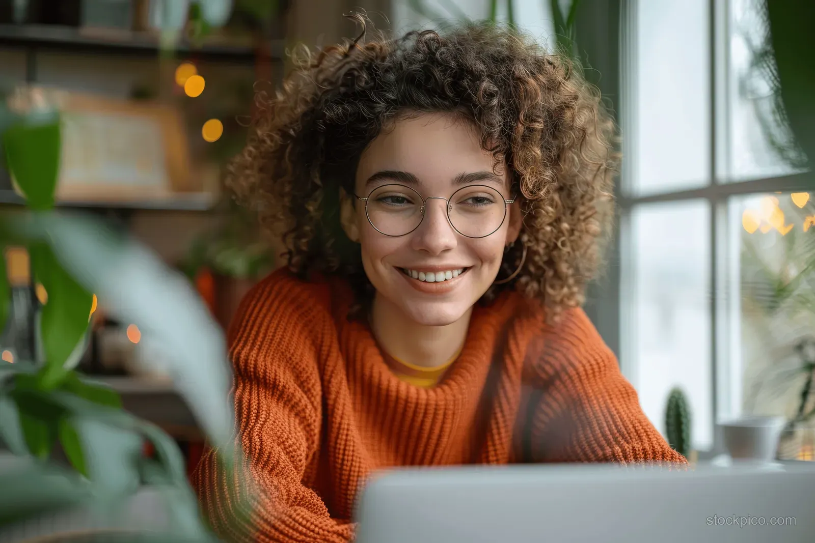 A smiling woman in glasses takes notes by her laptop during a virtual webinar in a plant-filled home office.