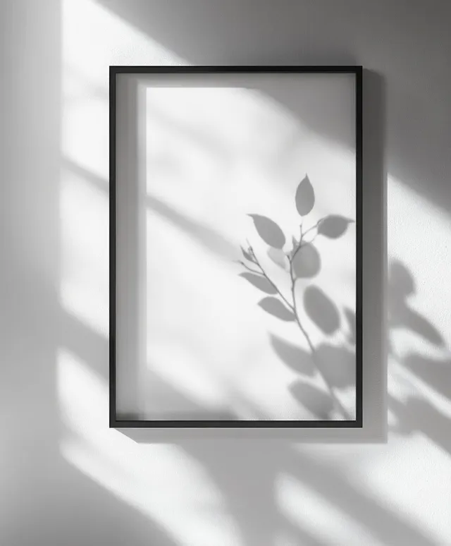 Minimalist black poster frame on a concrete wall with a blank insert and leaf-shaped shadows from sunlight.