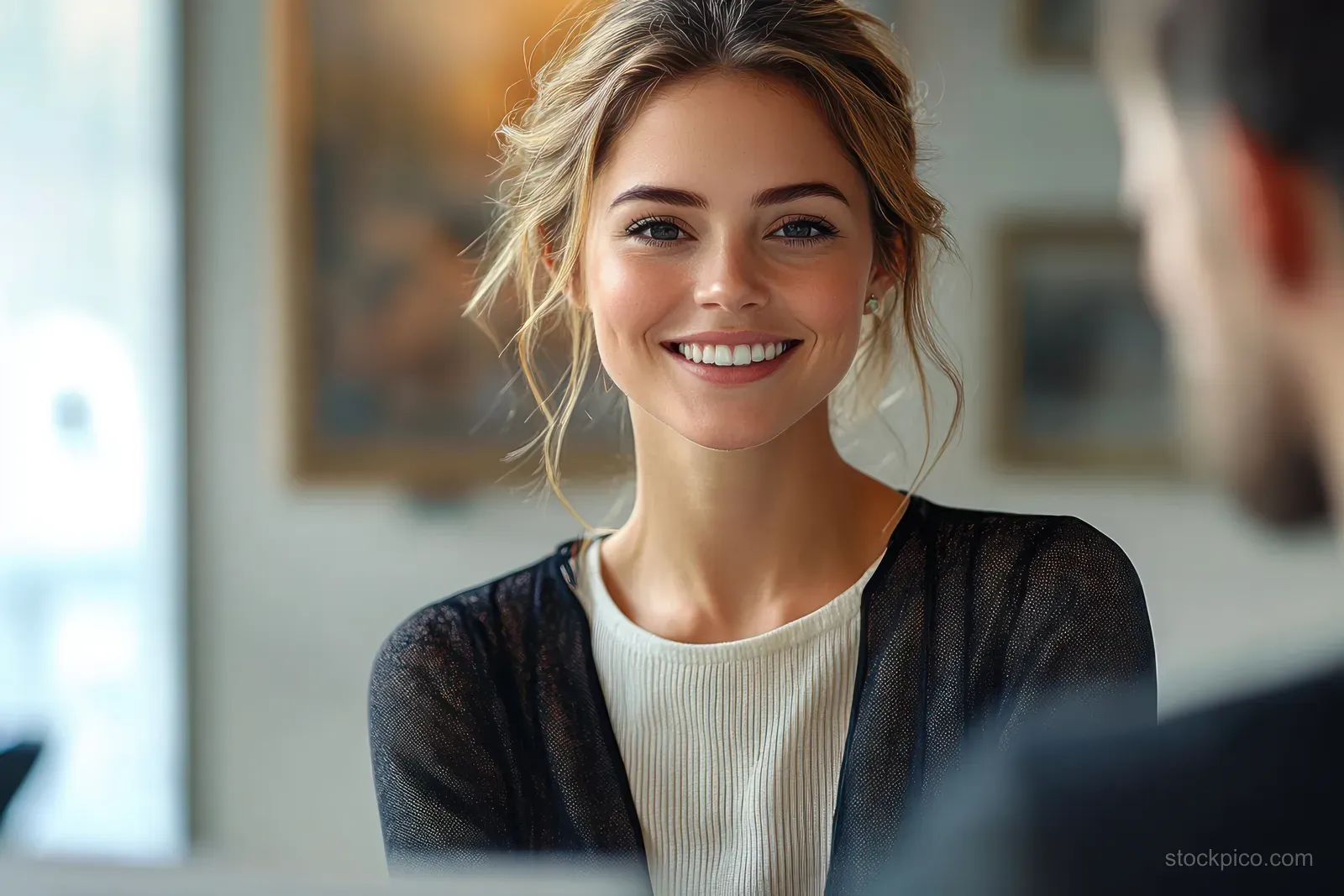 A smiling blonde woman in a black cardigan over a white top looks toward someone indoors in warm light.