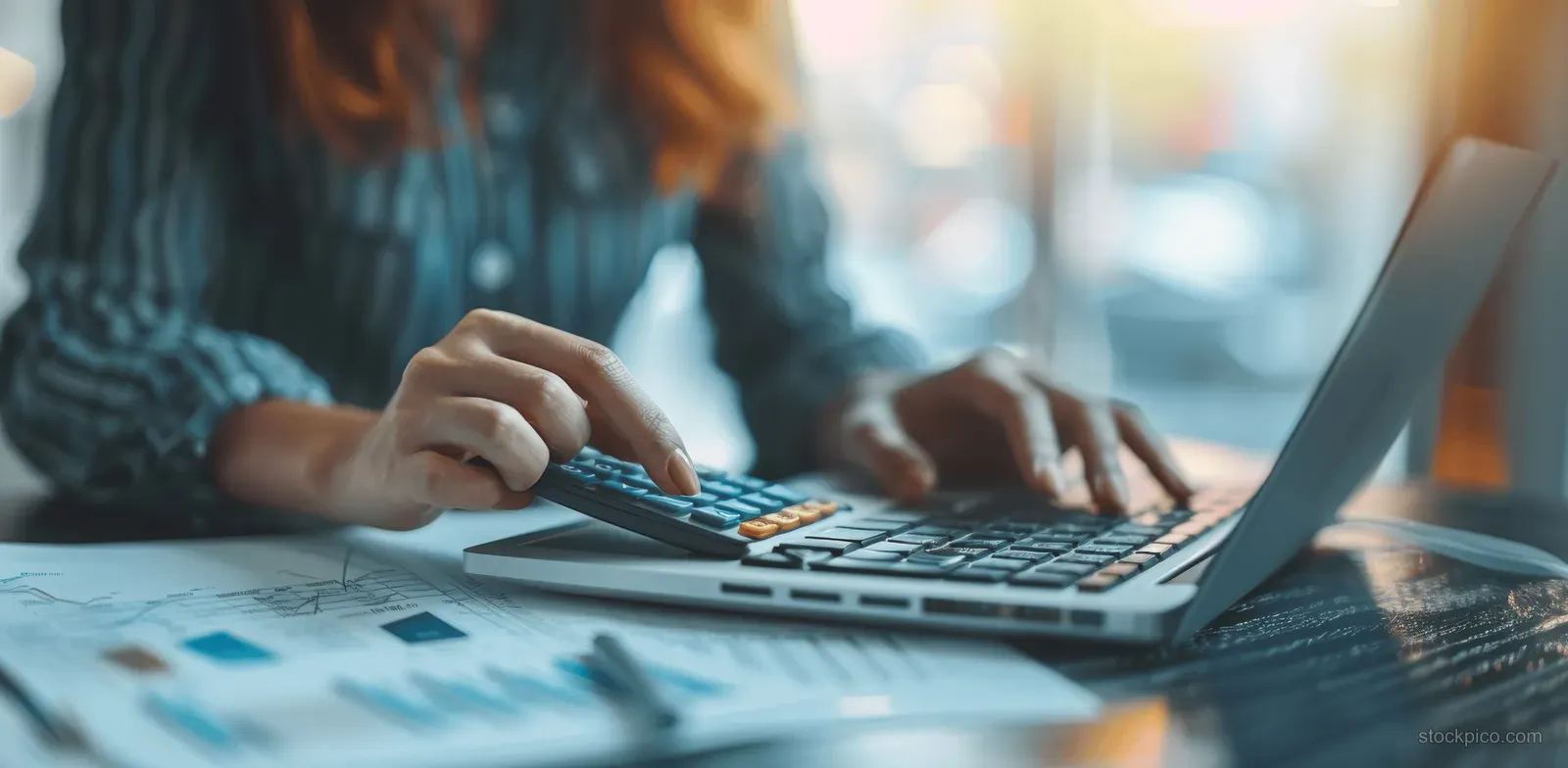 Businesswoman balances budget calculations with a calculator while reviewing figures on a laptop at her desk.