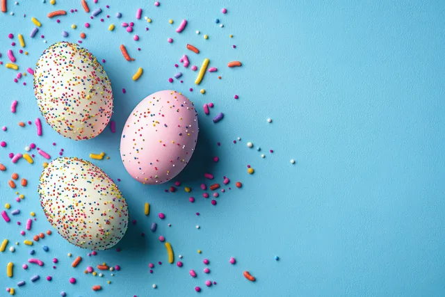 Three Easter eggs with multicolored sprinkles rest on a bright blue surface with scattered sprinkles.
