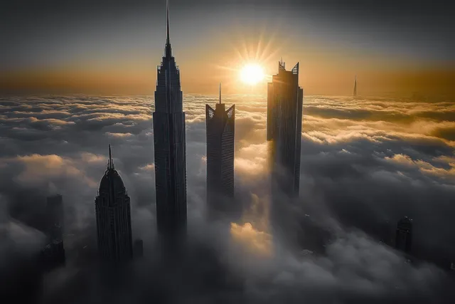Sunrise shines behind skyscrapers above thick fog and low clouds, creating golden sun rays and dark building silhouettes.