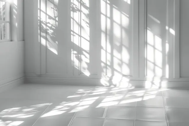Sunlight grid through a window casts long shadows over a white tiled floor and wall in a minimalist room.