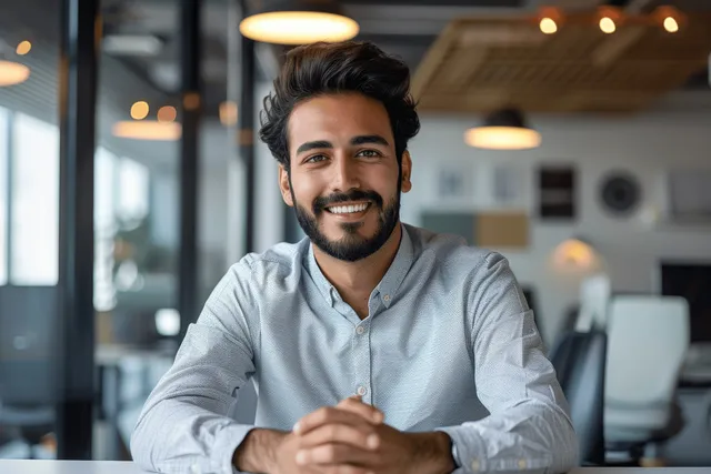 Smiling businessman sitting at a modern office desk, facing the camera with hands clasped.
