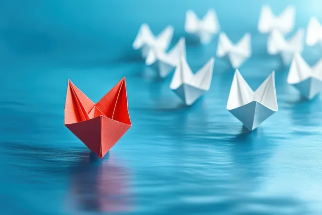 Red paper boat leads away from white paper boats across blue water with Creative Problem Solving
