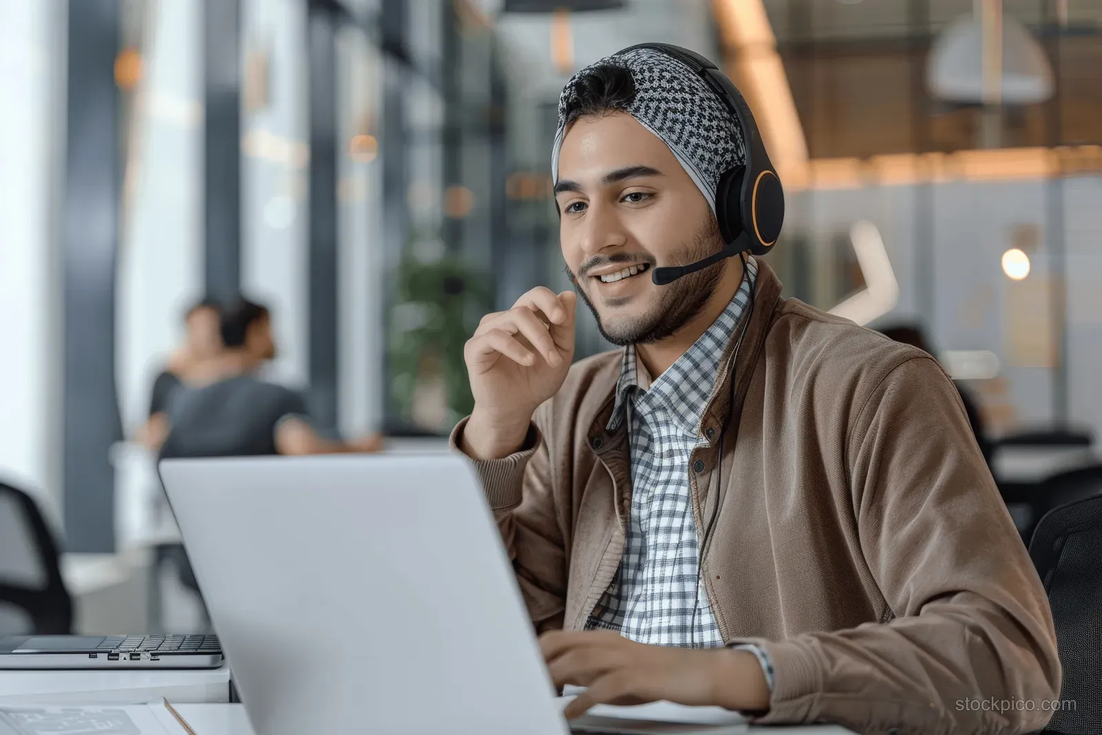 Cheerful young man wearing headphones speaks on a video call at a laptop in a modern office.