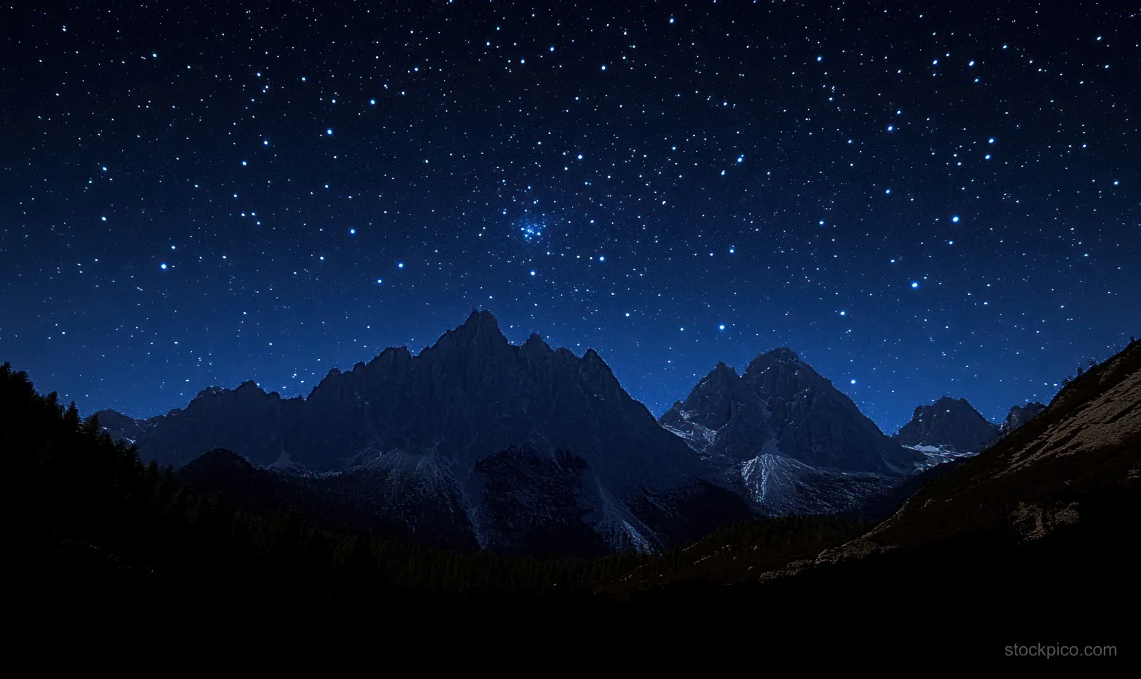 Countless stars illuminate jagged mountain peaks above a dark forest valley at night.