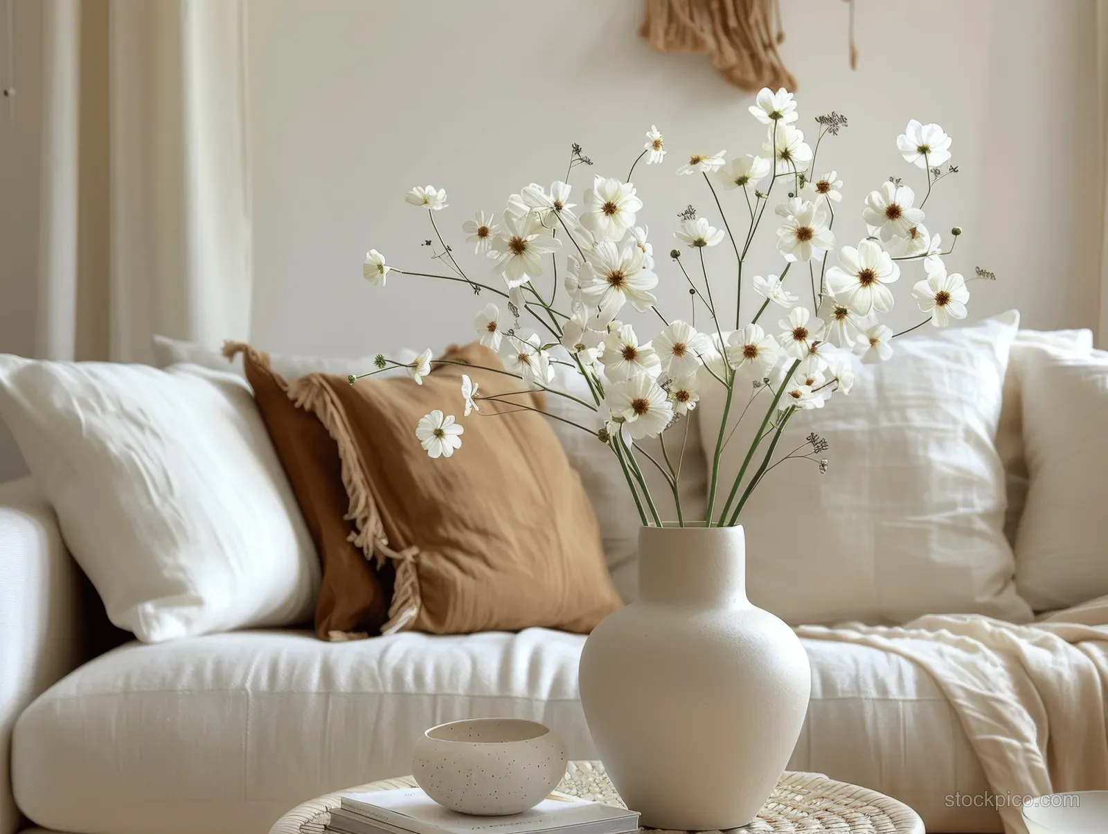 Beige ceramic vase with white flowers on a coffee table in front of beige sofa pillows.