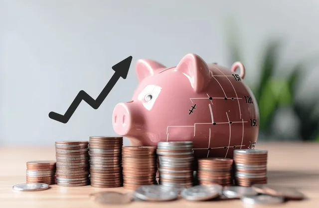 Pink piggy bank beside stacked coins with a black upward arrow indicating savings growth on a desk.