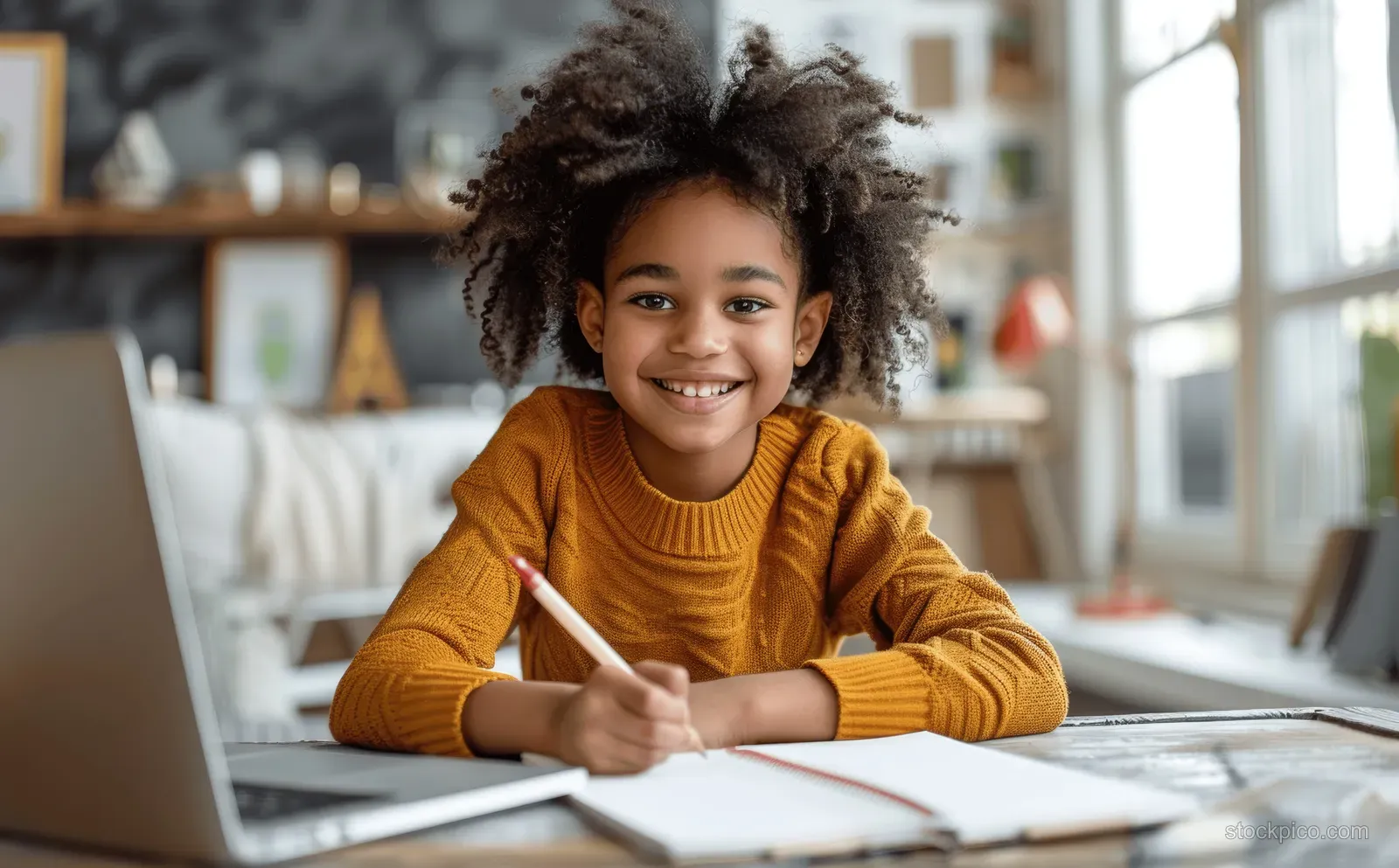 A smiling African American child writes homework at home while looking at an open laptop on the table.