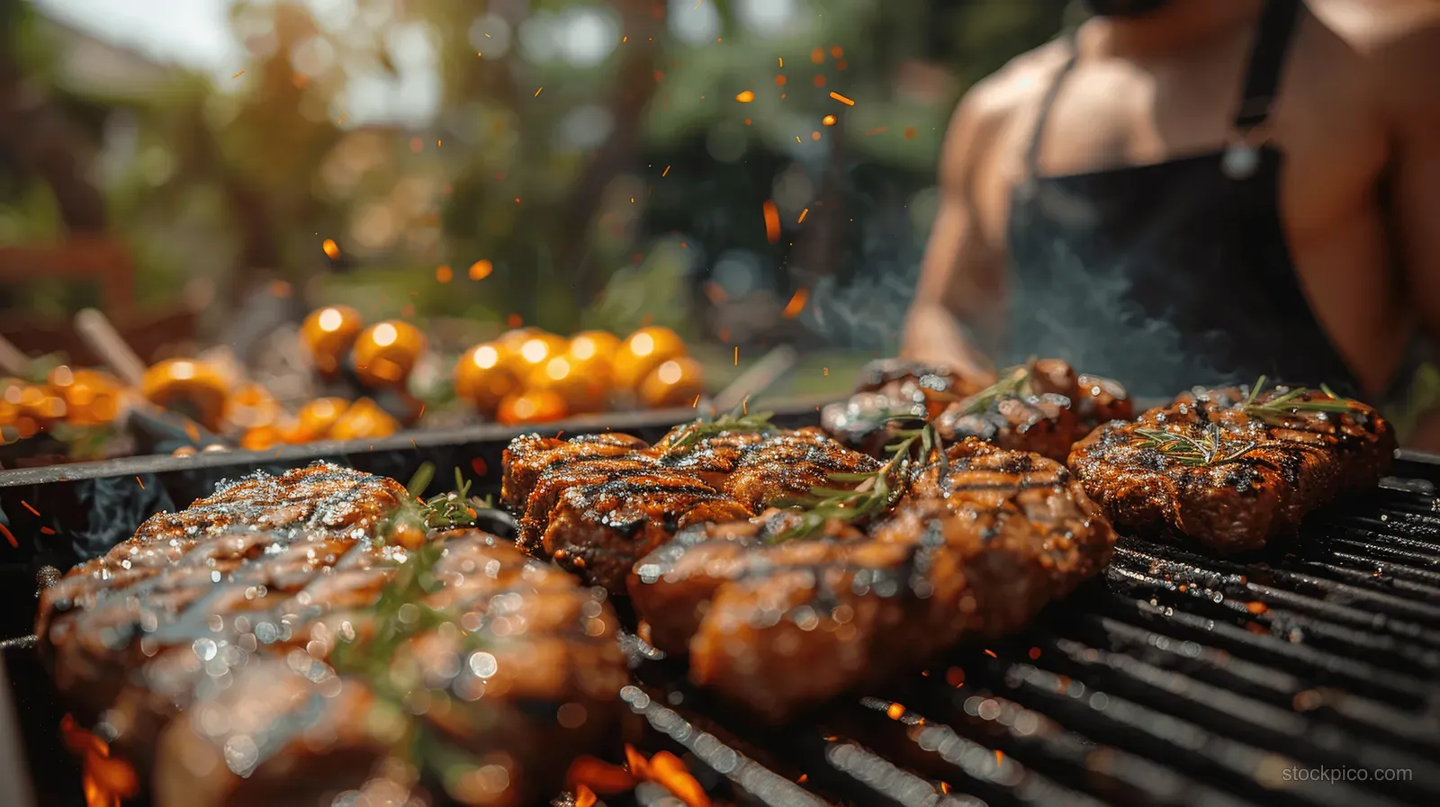 Close-up of steaks and sausage grilling on a hot barbecue grate in a backyard with people blurred behind.