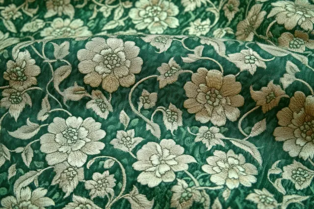 Textured green fabric with beige floral embroidery of stitched flowers and swirling stems fills the frame.