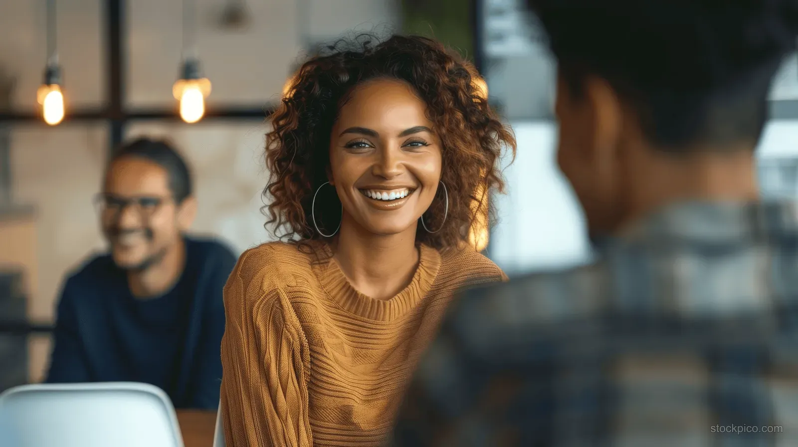 A smiling woman with curly hair sits at a cafe table while people are blurred in the background.