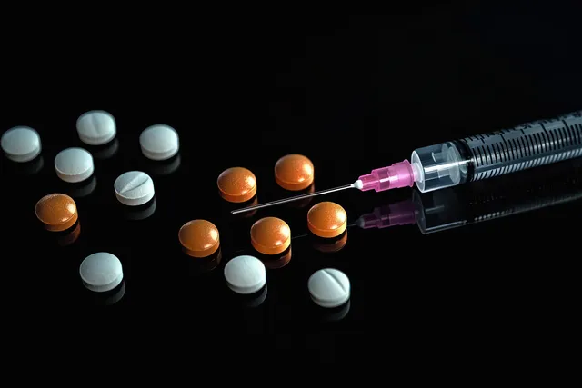 A syringe with pink needle cap lies next to scattered white and orange pills on a reflective black background.