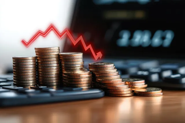 Stacked coins on a desk with a red downward chart line over a blurred laptop market screen.