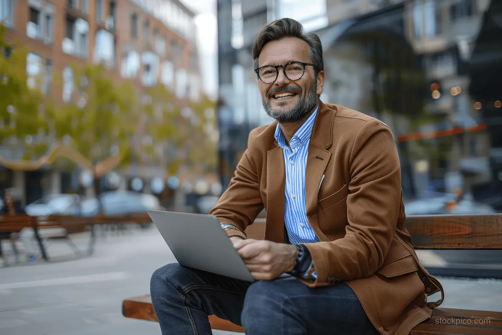 Smiling businessman in brown suit sits on an outdoor bench using a laptop and talking on a phone.