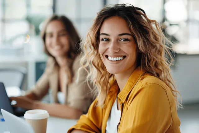 Two business women are smiling and collaborating during a meeting in a bright office with laptops and coffee cups.
