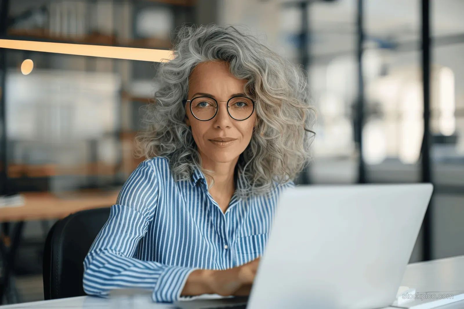 Mature woman executive with glasses typing on a laptop at a desk in a modern office.