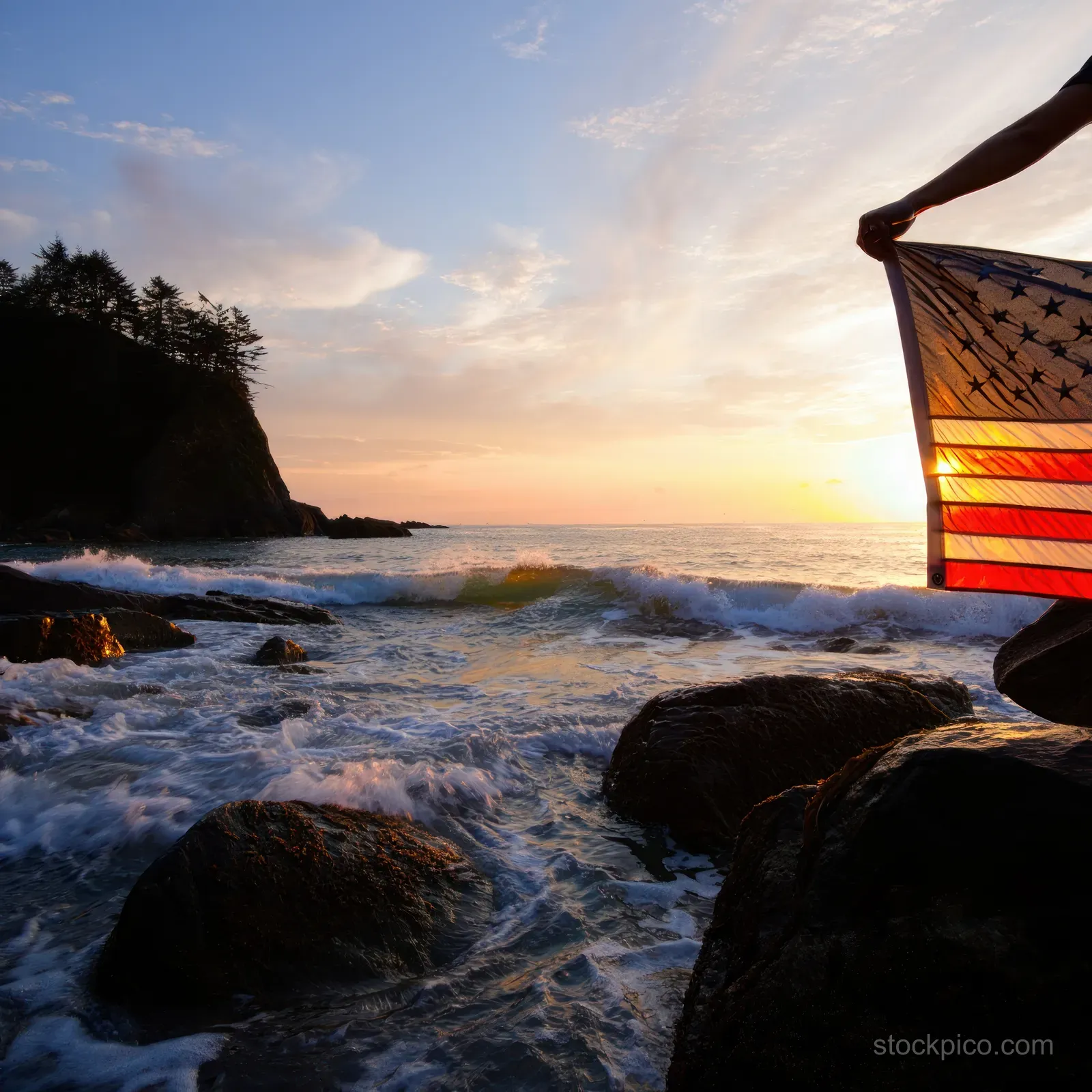 A person holds a partially transparent American flag at a rocky coastline as waves crash during a colorful sunset.